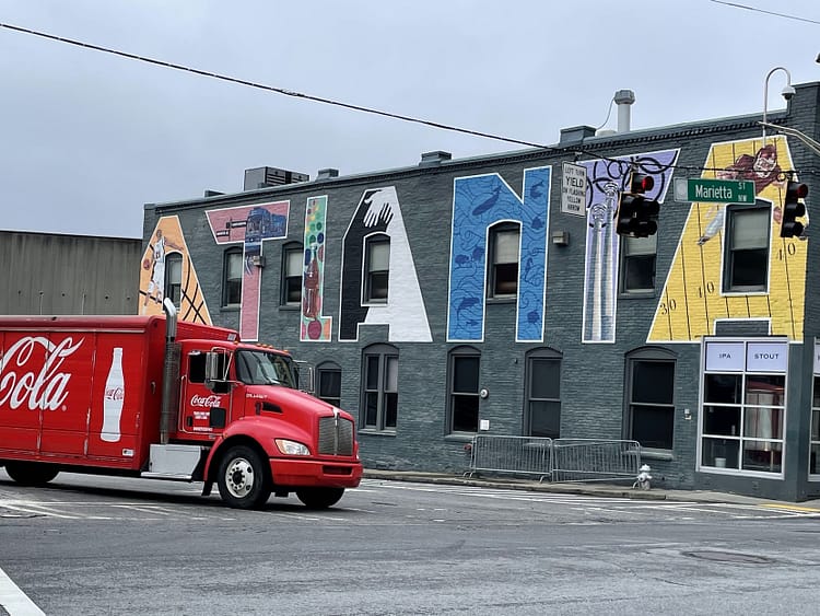 A Red Coca Cola delivery truck turned a corner in front of a gray building with Atlanta painted in giant block-style letters. Letter features a different attraction.