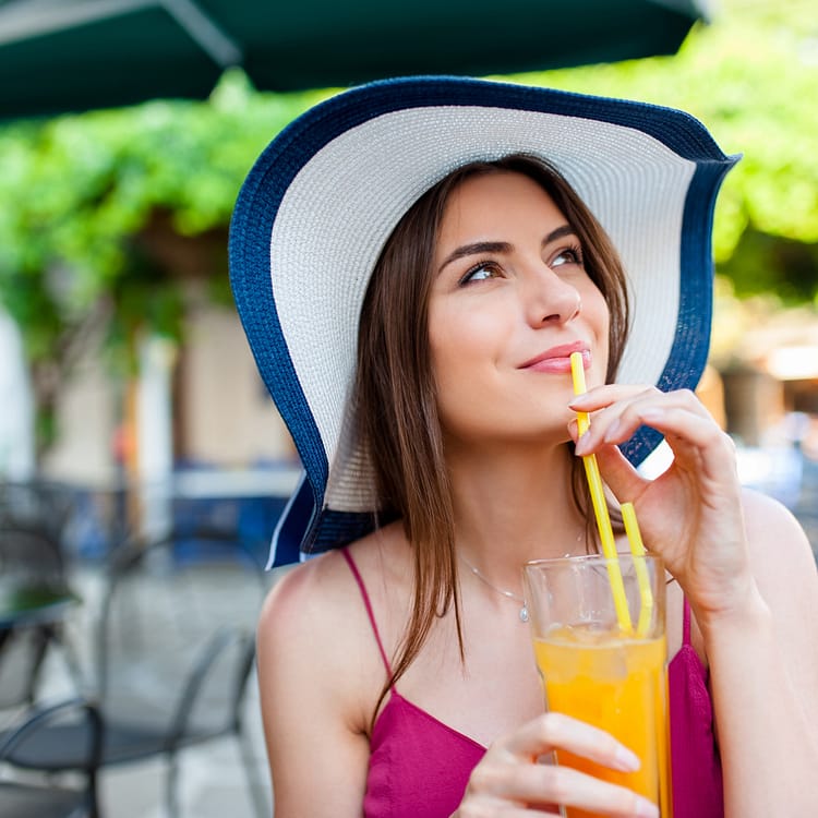 woman wearing a white sun hat trimmed in blue sitting at a table drinking an orange drink
