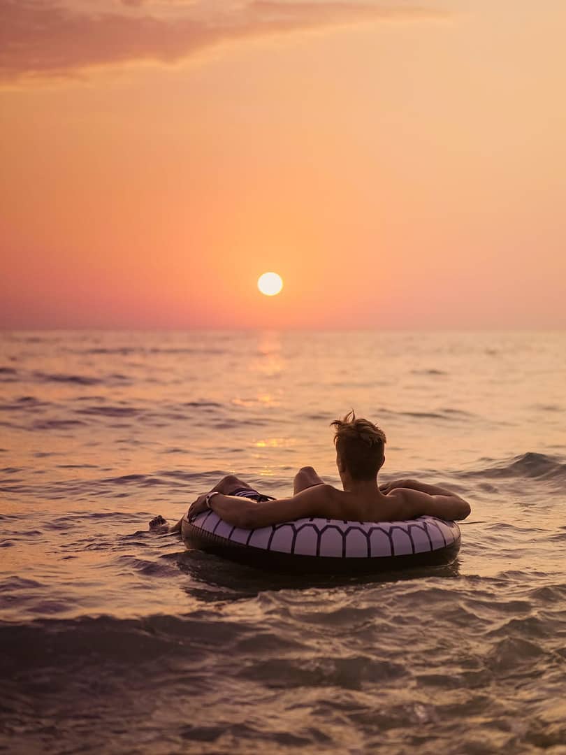 faceless male traveler in inflatable ring on ocean admiring sunset