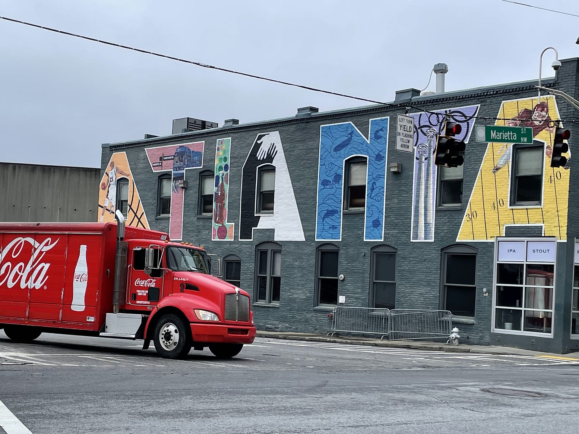 A Red Coca Cola delivery truck turned a corner in front of a gray building with Atlanta painted in giant block-style letters.  Letter features a different attraction.
